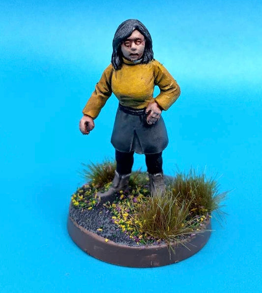 A painted tabletop miniature figurine of a woman with dark hair, wearing a mustard yellow top and dark skirt, standing on a circular base with grass and small flowers against a bright blue background.